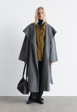 Übergroßer grauer Wollmantel mit breiten Revers über einem grünen Hemd mit Knöpfen, schwarzen Hosen und einer braunen Handtasche. Glatte Textur, lässige Passform.