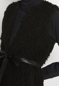 Czarna, teksturowana kamizelka z włochatym materiałem, o dopasowanej sylwetce, z czarną satynową wstążką zawiązaną w talii nad ciemnym topem.