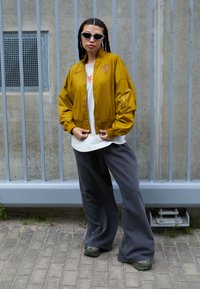 Jachetă de tip bomber aurie cu manșete ribbate, tricou alb, pantaloni largi gri și pantofi sport verzi. Ochelari de soare cu un design cat-eye.