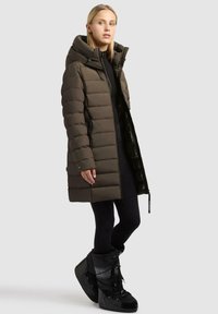 Vadderad, mörk olivgrön parka med hög krage, dragkedja och sidofickor; ihop med svarta isolerade stövlar och leggings.