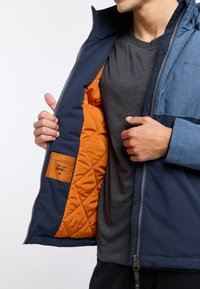 Giacca da uomo imbottita con esterno blu navy, pannello azzurro chiaro e foderatura trapuntata arancione vivace con etichetta del marchio all'interno.