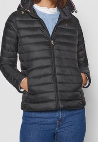 Černá péřová bunda s předním zipem, horizontálním prošíváním, kapucí s kontrastním lemem a zlatými doplňky. Nosená přes světle modrý svetr.