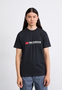 Sort bomulds T-shirt med "new balance" logo i hvid og rød, rund hals og korte ærmer, parret med grå bukser.