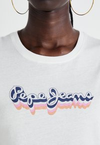 Close-up of a person wearing gold hoop earrings and a white t-shirt with "Pepe Jeans" written in navy, pink, and orange retro-style script.