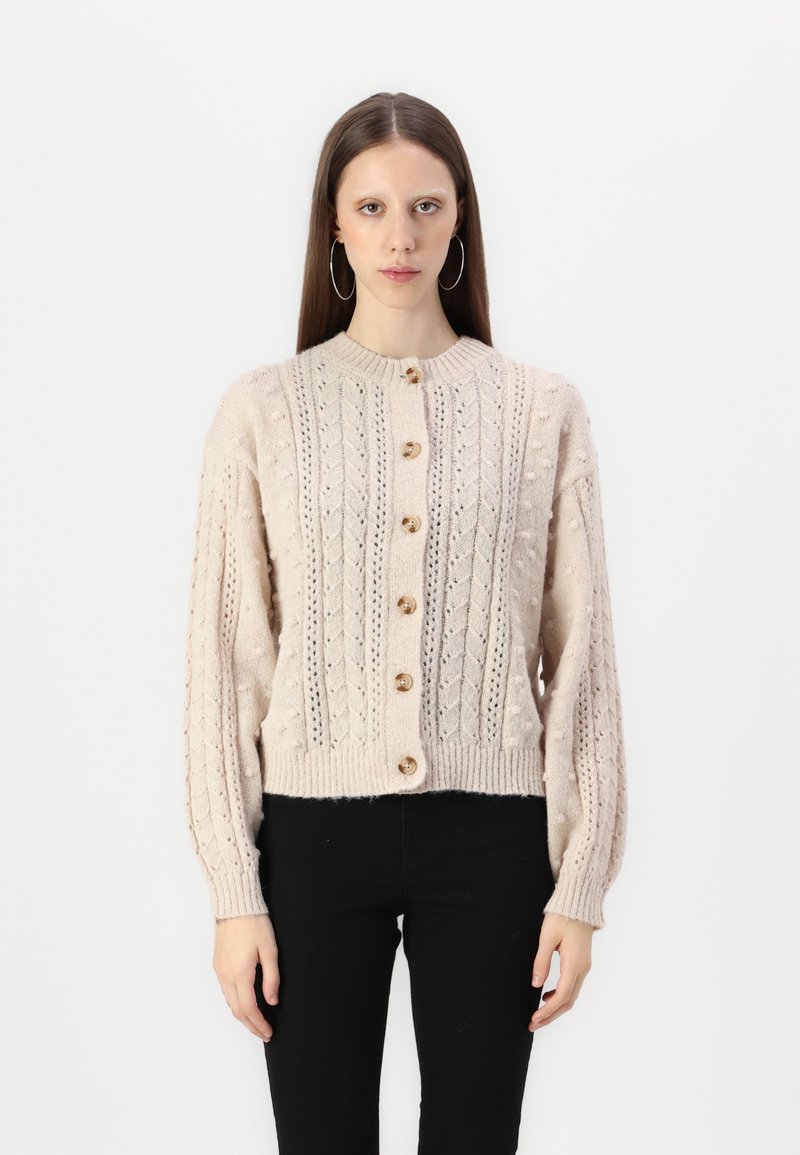 Cárdigan de punto color crema con cuello redondo, patrones texturizados y botones de madera en la parte delantera; mangas largas y abullonadas que se ajustan en los puños.