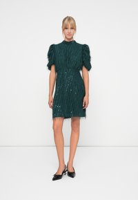 Green sequin dress with puff sleeves and fringed hem, crafted from textured material. Paired with black bow-detail heels.