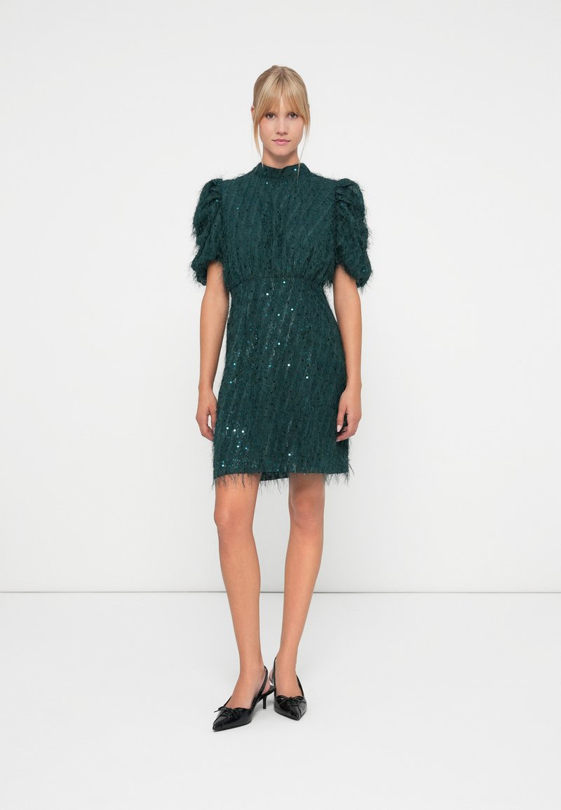 Green sequin dress with puff sleeves and fringed hem, crafted from textured material. Paired with black bow-detail heels.