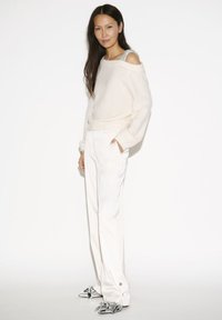 Vrouw met lang, donker haar in een off-shoulder crèmekleurige trui, witte brede broek en zilveren puntige schoenen, staande met handen in de zakken.