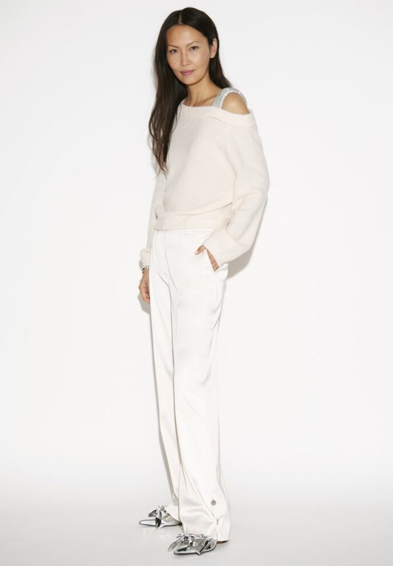 Vrouw met lang, donker haar in een off-shoulder crèmekleurige trui, witte brede broek en zilveren puntige schoenen, staande met handen in de zakken.