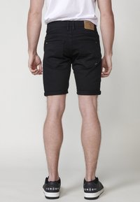 Muž v čiernych skrátených džínsach, bielom tričku a čiernych teniskách s bielymi podrážkami stojí pred jednoduchým svetlým pozadím.