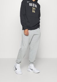 Sweat à capuche Nike noir avec motif doré et blanc ; pantalon de survêtement gris ; chaussures de sport blanches. Le sweat à capuche comporte une poche avant et une cagoule à cordon.
