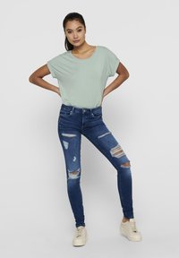 Mintgrünes T-Shirt mit kurzen Ärmeln, kombiniert mit verwaschenen blauen Skinny-Jeans. Weiße Plateausneaker runden das Outfit ab.