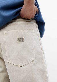 Mano metida en el bolsillo de unos pantalones vaqueros beige de Pull&Bear, combinados con una camiseta azul, contra un fondo blanco.