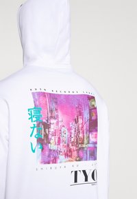 Sweat à capuche blanc avec un graphisme néon coloré représentant le paysage urbain de Shibuya à Tokyo et un texte en japonais au dos.