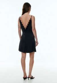 Schwarzes Satin-Kleid mit tiefem V-ausschnitt, verziert mit Spitzenbesatz entlang des Ausschnitts und einem ausgestellten Saum. Dünne, verstellbare Träger, knielang.