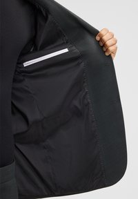Zwarte blazer binnenkant met een gladde, glanzende voering, een witte zak en zorgvuldig genaaide naden. De buitenkant is van een gestructureerde donkere stof.