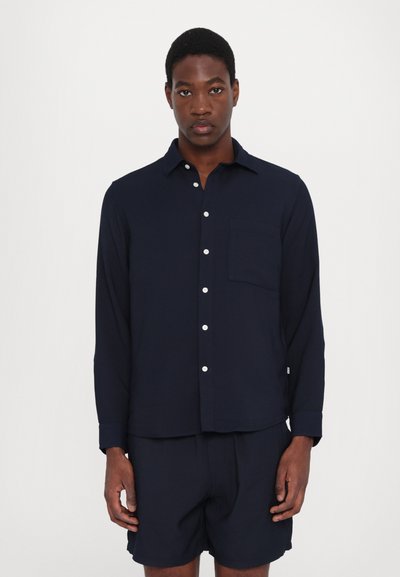 Navyblaues Langarmhemd mit durchgehender Knopfleiste, Brusttasche und abgerundetem Saum, kombiniert mit passender Shorts in einem ähnlichen Farbton.