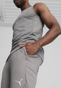 Grijze sportieve tanktop met een gestructureerd ontwerp en bijpassende grijze broek met een reflecterende Puma-logo. De stof lijkt licht en soepel.