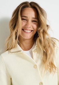 Cardigan de color crema claro con cuello texturizado y detalles dorados en los botones, hecho de una tela de punto con un patrón de canalé.