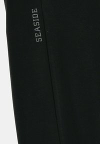 Tissu noir avec texte vertical gris "SEASIDE" imprimé près du bord, montrant un détail de couture subtil.