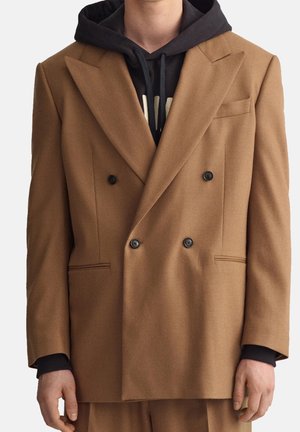 Mężczyzna w dwurzędowej, wielbłądziej marynarce noszonej na czarnej bluzie z widocznym kapturem i sznurkami, stojący na jednolitym tle.