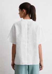 Weißes Leinenoberteil mit kurzen Ärmeln, rückseitiger Nahtdetails und einem runden Kragen. Kombiniert mit hellblauen, locker sitzenden Hosen.