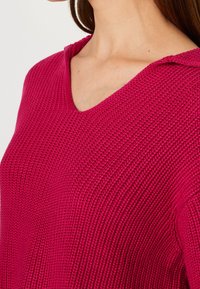 Femme portant un pull en maille texturée rose profond avec un col en V, se concentrant sur la partie supérieure de la poitrine et des épaules.
