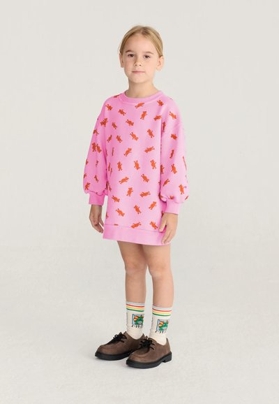 Jeune fille portant une robe rose avec des imprimés d’ours en peluche orange, des chaussettes colorées à motifs et des chaussures marron à lacets, debout sur un fond clair.