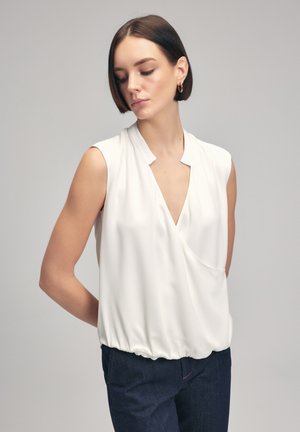 Blusa bianca senza maniche con scollo a V e parte anteriore drappeggiata, caratterizzata da un orlo arricciato. Tessuto morbido e leggero. Indossata con jeans blu scuro.