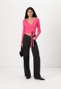 Har en rosa stripete langermet innhyllet topp med knyting i midjen, kombinert med høyt liv svarte vide jeans og svarte spisse sko.