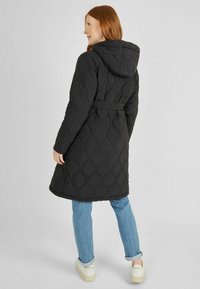 Abrigo largo acolchado negro con capucha, cintura con cinturón y patrón ondulado texturizado, combinado con jeans azul claro y zapatillas blancas.