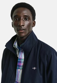 Giacca navy con colletto alto, indossata sopra una camicia a quadri multicolore con strisce blu, verdi e viola. Logo sul petto.