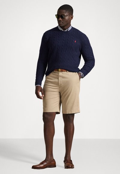 Ναυτικό πουλόβερ με πλέξη κασμίρι, ανοιχτό μπλε πουκάμισο με γιακά, καφέ σορτς και καφέ loafers. Τα αξεσουάρ περιλαμβάνουν σκούρα γυαλιά ηλίου και καφέ ζώνη.