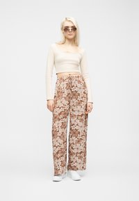 Γυναίκα που φοράει μπεζ μακρυμάνικο crop top, καφέ φαρδιά παντελόνια με floral σχέδιο, λευκά αθλητικά παπούτσια και γυαλιά ηλίου, στέκεται μπροστά σε απλό λευκό φόντο.