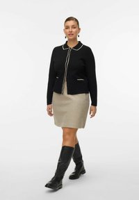 Kobieta idąca w czarnym kardiganie zapinanym na guziki z białymi wykończeniami, beżowej spódnicy do kolan, czarnych kozakach do kolan oraz kolczykach fasonu drop.