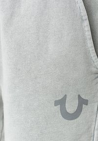 Světle šedé šortky vyrobené z měkké tkaniny, které mají hladký povrch a výrazné tmavě šedé logo na levé straně.