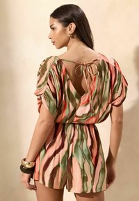 Mujer con cabello largo y oscuro lleva un mono multicolor a rayas en rosa, verde y marrón con una espalda profundamente abierta atada con un lazo y pulseras gruesas.
