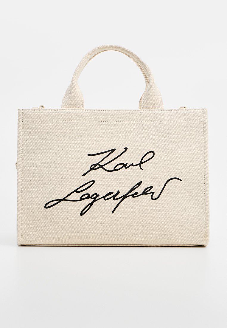 Sac fourre-tout rectangulaire beige en toile, avec des poignées courtes et un script cursif noir centré sur le devant.