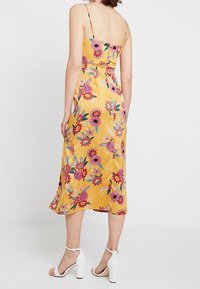 Gelbes Blumenkleid mit dünnen Trägern, hat ein enganliegendes Oberteil und einen fließenden Midi-Rock. Der Stoff hat eine glatte Textur, geschmückt mit bunten Mustern.