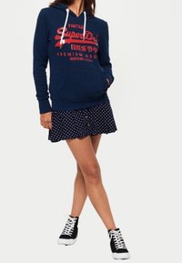 Mujer vestida con una sudadera SuperDry azul marino, una falda azul marino con lunares y zapatillas negras de caña alta con cordones blancos, de pie con las piernas cruzadas.