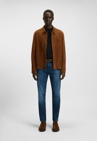 Brązowa zamszowa kurtka z zapięciem na guziki, dwie przednie kieszenie, czarny t-shirt z okrągłym dekoltem, niebieskie jeansy slim-fit, brązowe botki za kostkę. Gładkie tło.