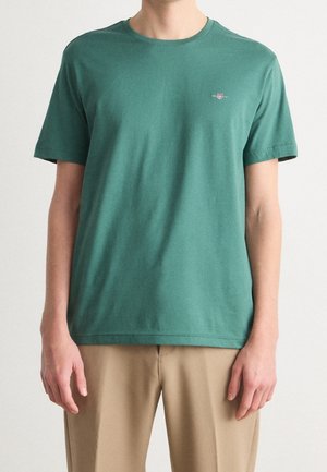 Homme portant un t-shirt vert uni à manches courtes avec un petit logo sur la poitrine et un pantalon beige, debout devant un fond uni.