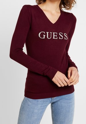 Kobieta ubrana w dopasowany bordowy sweter z dekoltem w serek z napisem „GUESS” z cyrkonii oraz jasnoniebieskie jeansy, ręce splecione z przodu.