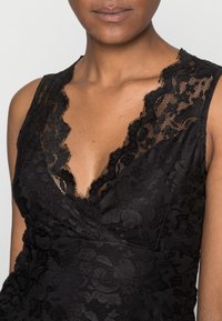 Donna che indossa un vestito nero senza maniche in pizzo con motivi floreali e una scollatura profonda a forma di V con bordi ondulati.