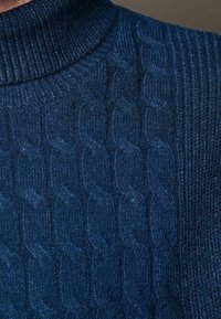 Gros plan d'un pull en maille torsadée bleu marine montrant des motifs texturés en tresses et un col côtelé.