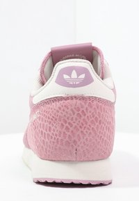 Rosa sneaker med ett texturerat, reptilliknande mönster, vita detaljer och en vadderad häl med en dovt rosa logotyp. Gummisula syns nedanför.