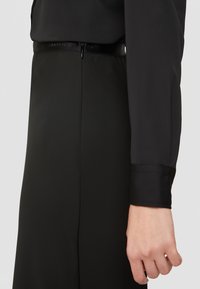 Černé hladké šaty z látky mají přiléhavý pas, jemný zip a saténové manžety. Minimalistický design bez vzorů nebo akcentů.