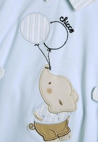Tessuto azzurro chiaro con un elefante ricamato che tiene in mano palloncini a righe nere e bianche. "Chicco" è ricamato in nero sopra il design.