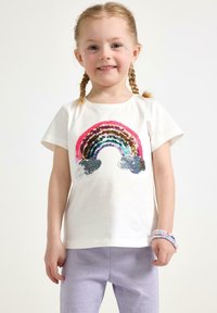 Vit bomulls-T-shirt med en flerfärgad paljettregnbågsdesign. Korta ärmar, bärs med lila stickade byxor och färgglada pärlarmband.