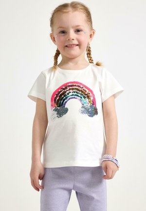Vit bomulls-T-shirt med en flerfärgad paljettregnbågsdesign. Korta ärmar, bärs med lila stickade byxor och färgglada pärlarmband.